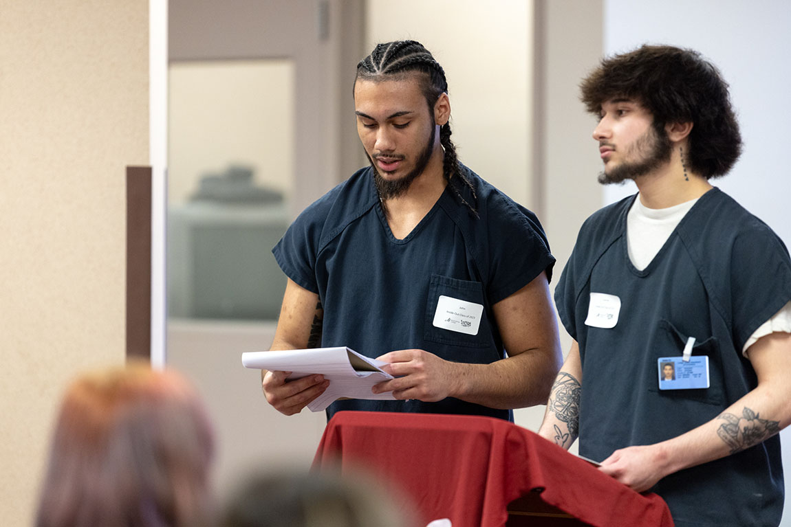 Two men in navy blue prison uniforms stand at a podium; the one on the left reads from a notebook